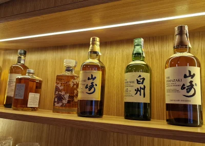Na obrázku je krásná sbírka prémiových japonských whisky, které jsou vystavené na podsvícené dřevěné polici. Skvěle to doplňuje luxusní atmosféru japonské restaurace