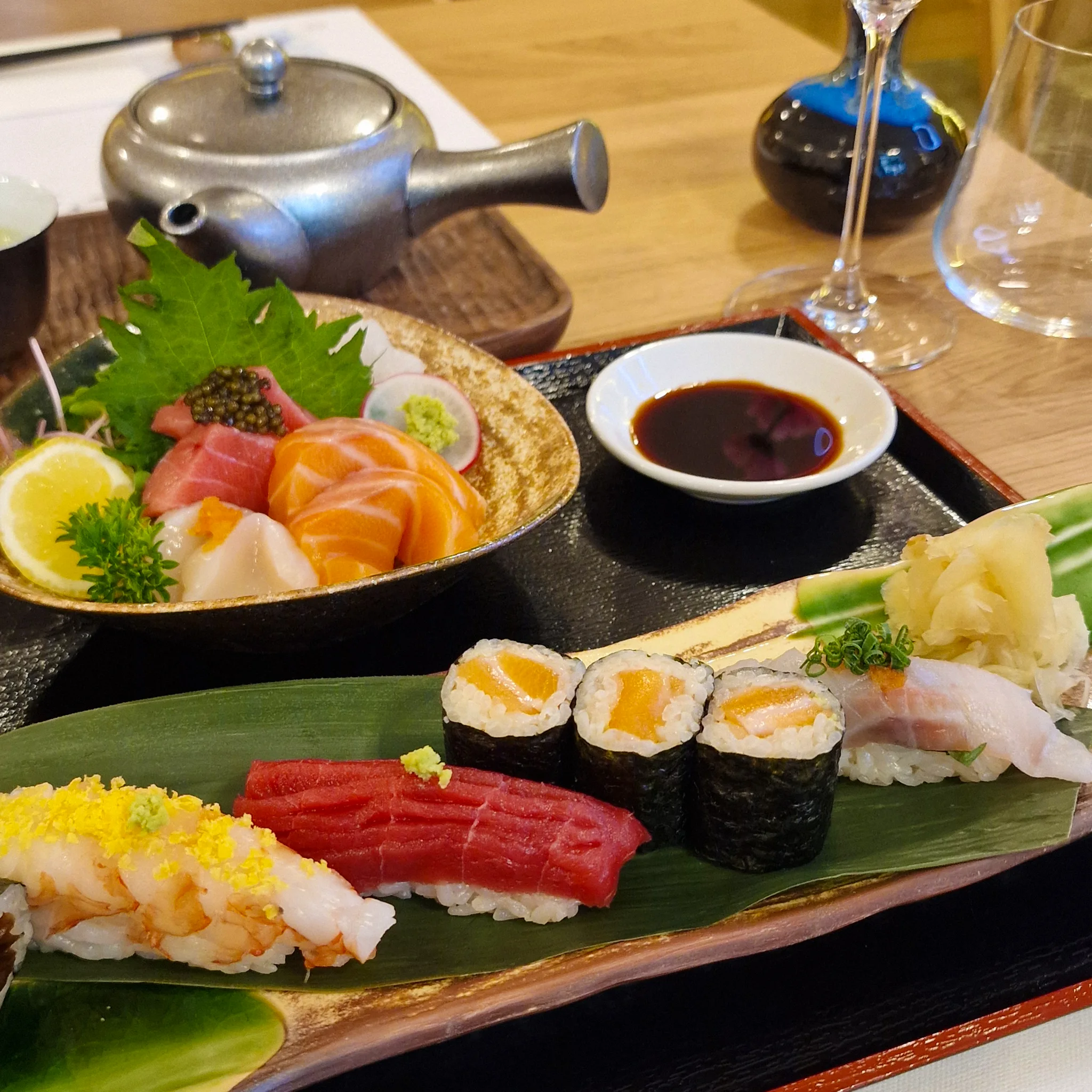 Image_rik8uerik8uerik8_0002_Vrstva-1 kopie 4 Japonské menu na černém podnosu. V popředí je podlouhlý talíř se sushi servírovaným na zeleném listu: nigiri s krevetou, nigiri s dlouhým plátkem tuňáka, tři maki rolky s lososem, nigiri s bílou rybou a hromádka nakládaného zázvoru.
